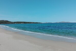 A beach with clear blue water, sandy shore, and distant hills.