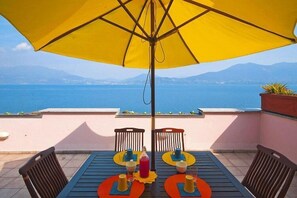 Une table entourée de chaises et surmontée d’un parasol jaune, sur une terrasse donnant sur une étendue d’eau, avec des montagnes en arrière-plan.