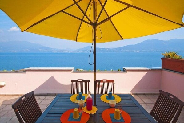 Una mesa con sillas y un paraguas amarillo en una terraza con vista a un cuerpo de agua y montañas al fondo.