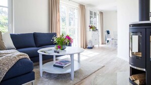 Ein Wohnzimmer mit einer Couch, einem runden Couchtisch mit Büchern und Blumen, einem Kamin und einem Fenster mit Vorhängen.