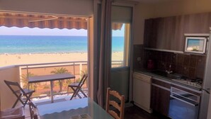 Un balcon avec vue sur la plage, une table à manger, des chaises et un coin cuisine équipé de placards et d’appareils électroménagers.