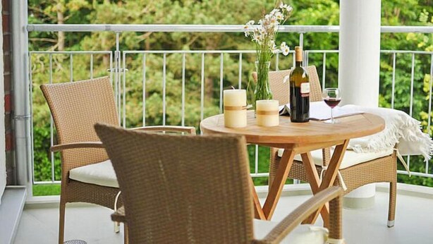 A balcony with wicker furniture, a round wooden table, candles, a bottle of wine, and a vase of flowers. - Wohnung in Cuxhaven. (Cuxhaven)