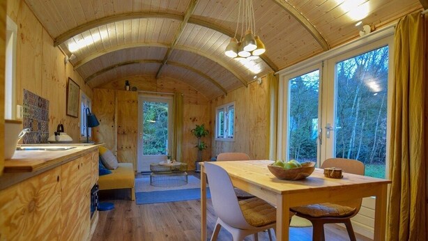 Interior de una cabaña de madera con comedor, cocina y ventanas amplias.