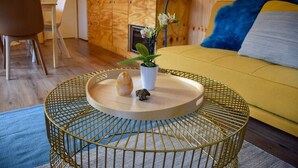 Una sala de estar acogedora con un sofá amarillo, una mesa redonda de hilo dorado con una bandeja, un jarrón blanco pequeño con flores y un cojín azul.