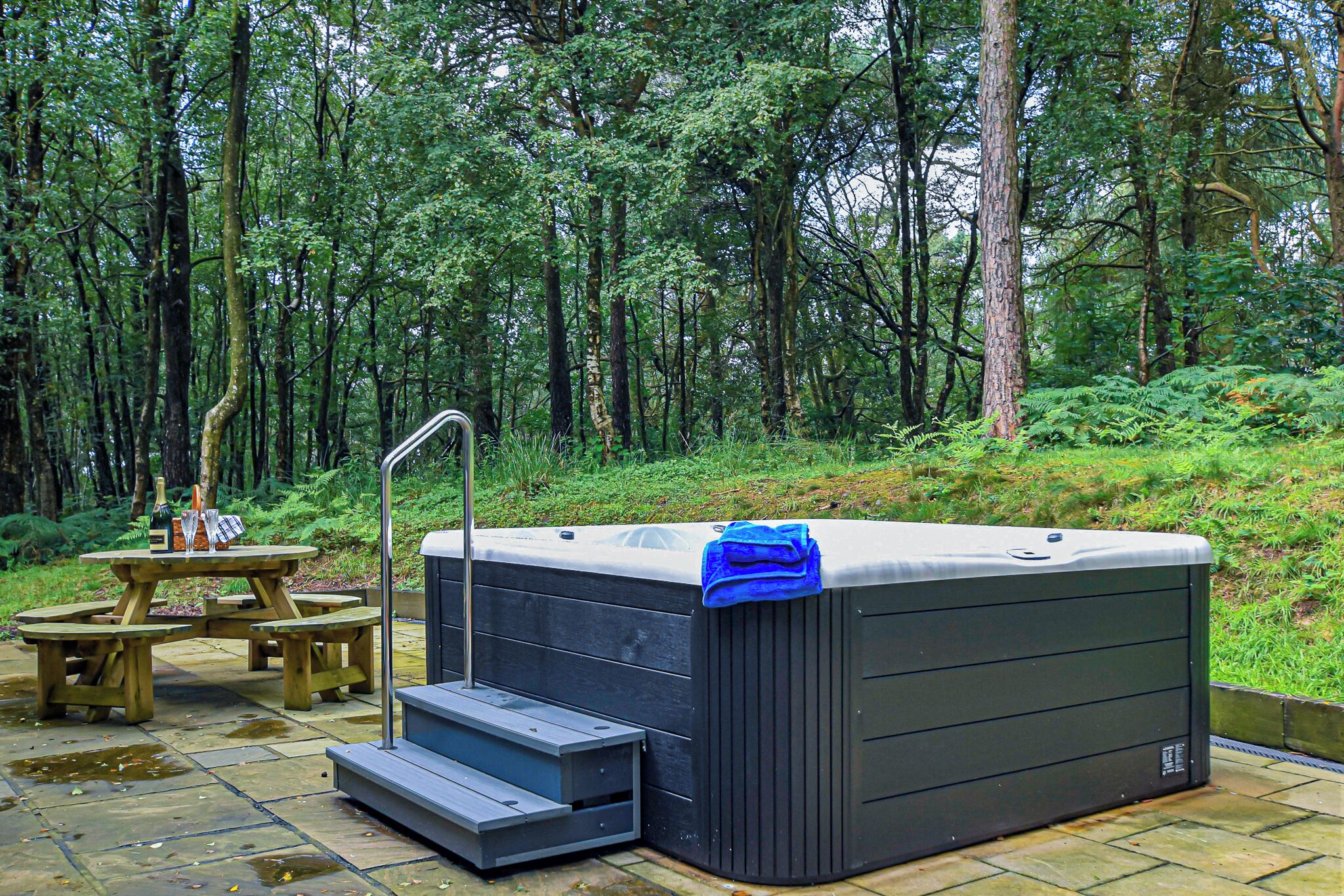 Un jacuzzi con una toalla azul, una mesa de picnic de madera y un patio de piedra en un entorno boscoso.