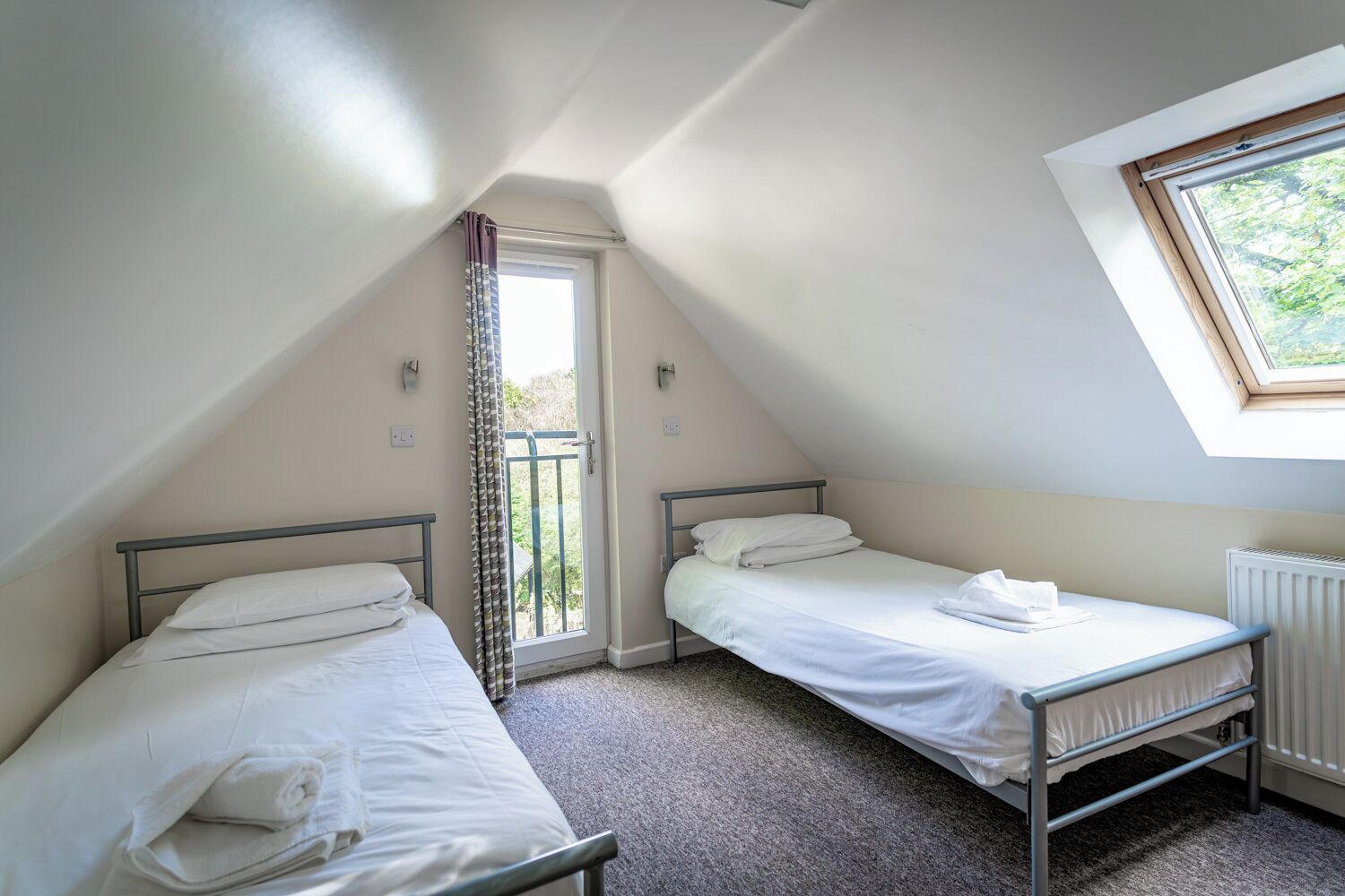 A small, well-lit room with two single beds, a window with a view of greenery, and a radiator.