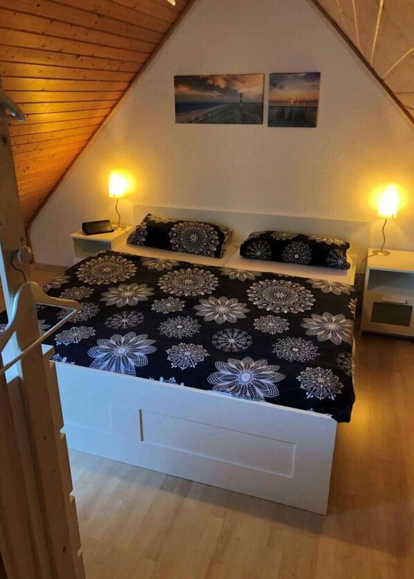 Ein Schlafzimmer mit schrägen Wänden, einem Bett mit floralem Überwurf, zwei Nachttischlampen und zwei gerahmten Bildern an der Wand.