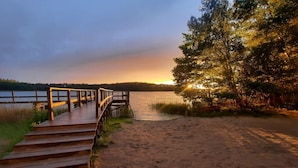 A wooden boardwalk leading to a sandy beach with a lake and trees in the background.