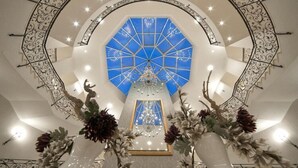 Een statig atrium met een groot blauw glas-in-loodraam, een centrale kroonluchter en decoratieve vazen met bloemen.