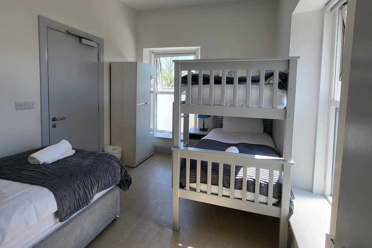 A compact room with a bunk bed, a single bed, a wardrobe, and a window providing natural light.