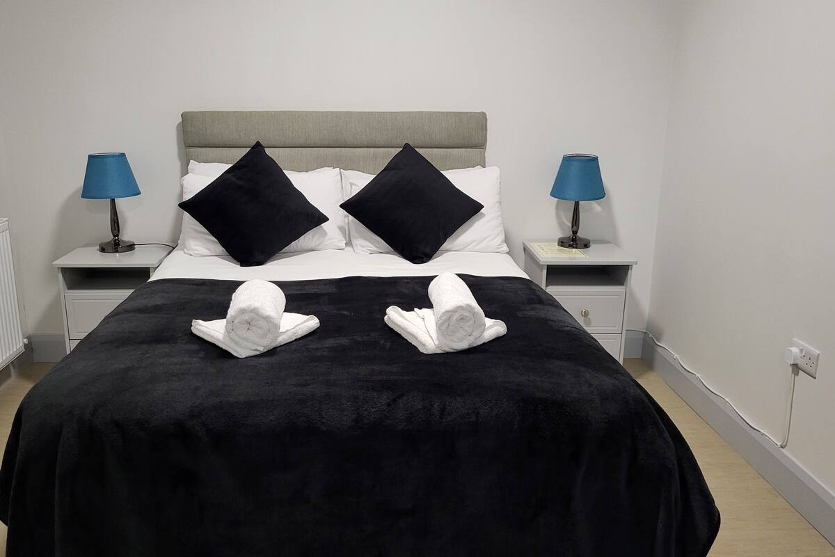 A neatly made bed with a black bedspread, two black pillows, and two rolled towels on the bed. There are two bedside tables with lamps and a white wall radiator.