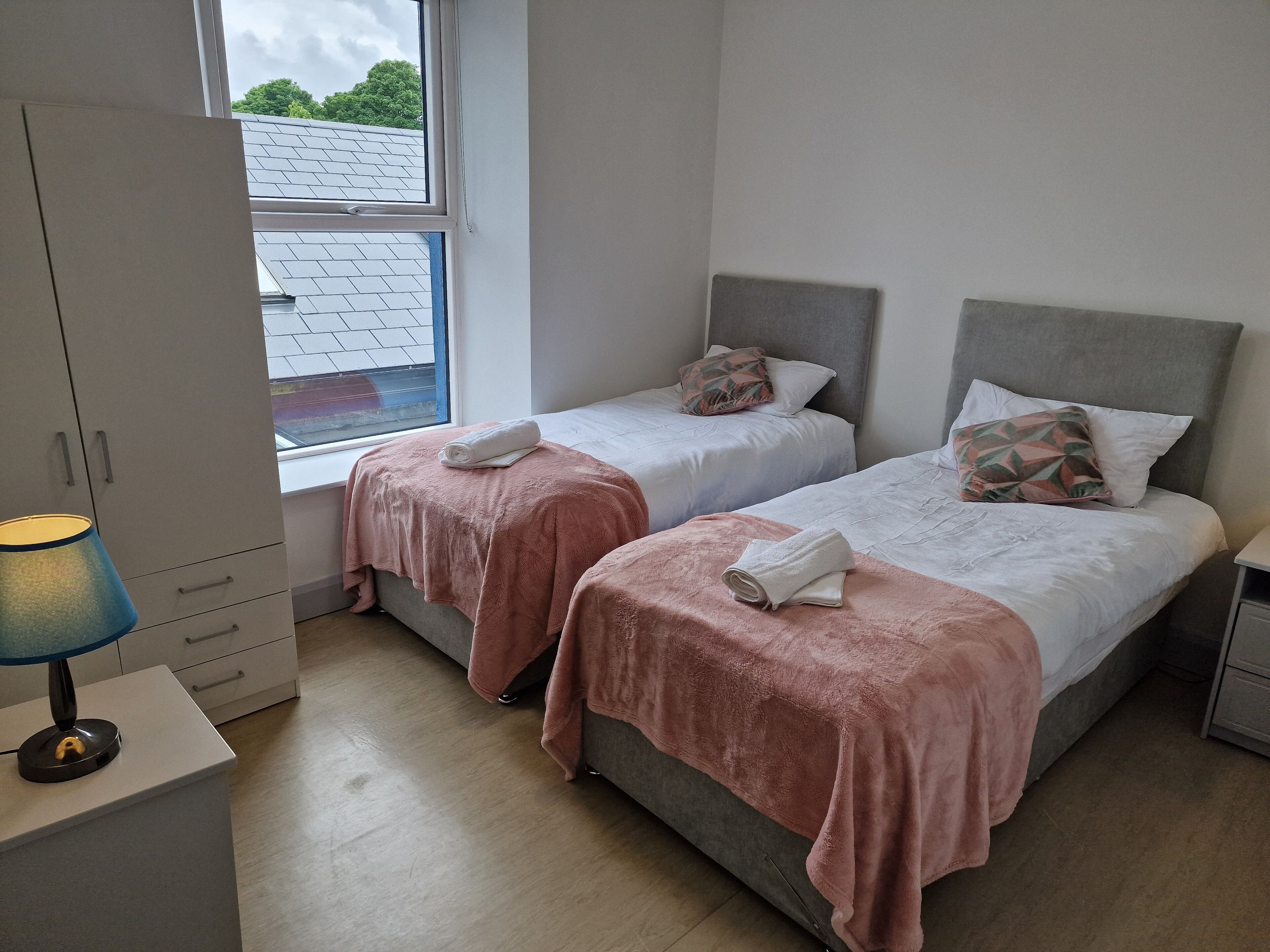 A bedroom with two beds, each with a pink blanket and pillows, a window with a view of a building, and a desk with a lamp.