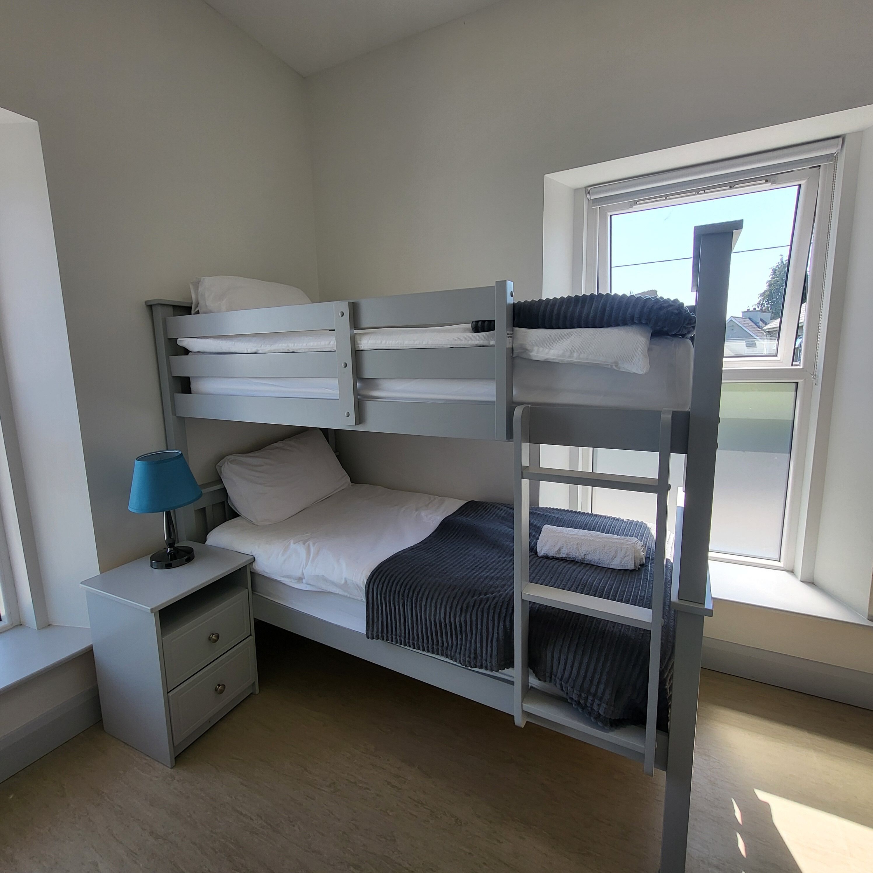Habitación con literas, escritorio, lámpara, ventana con vista a edificios y piso de madera.