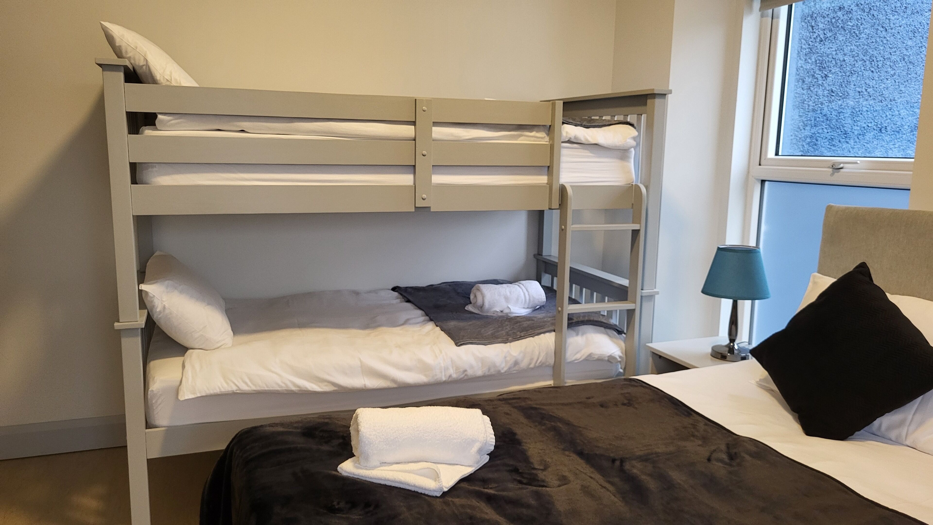 A bunk bed room with a window, a bedside table with a lamp, and a black pillow on the lower bunk.