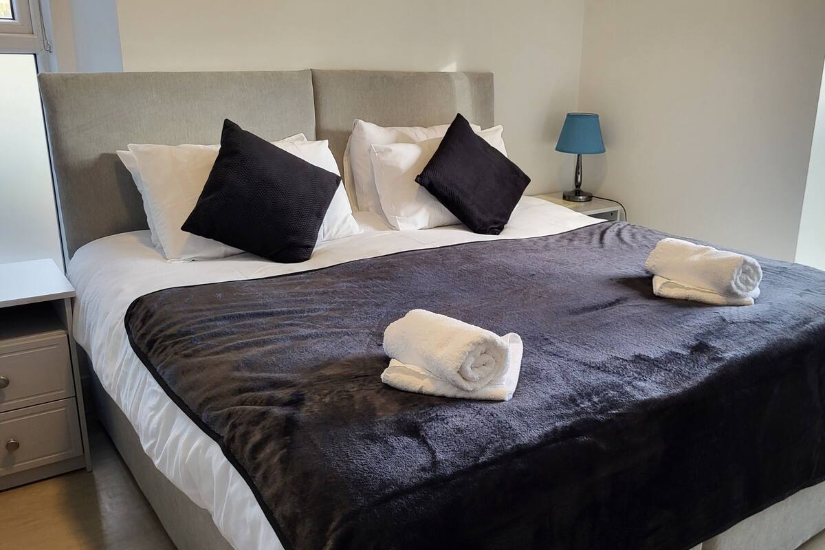 A neatly made bed with a dark blanket and two rolled towels, a bedside table with a lamp, and a window with blinds.