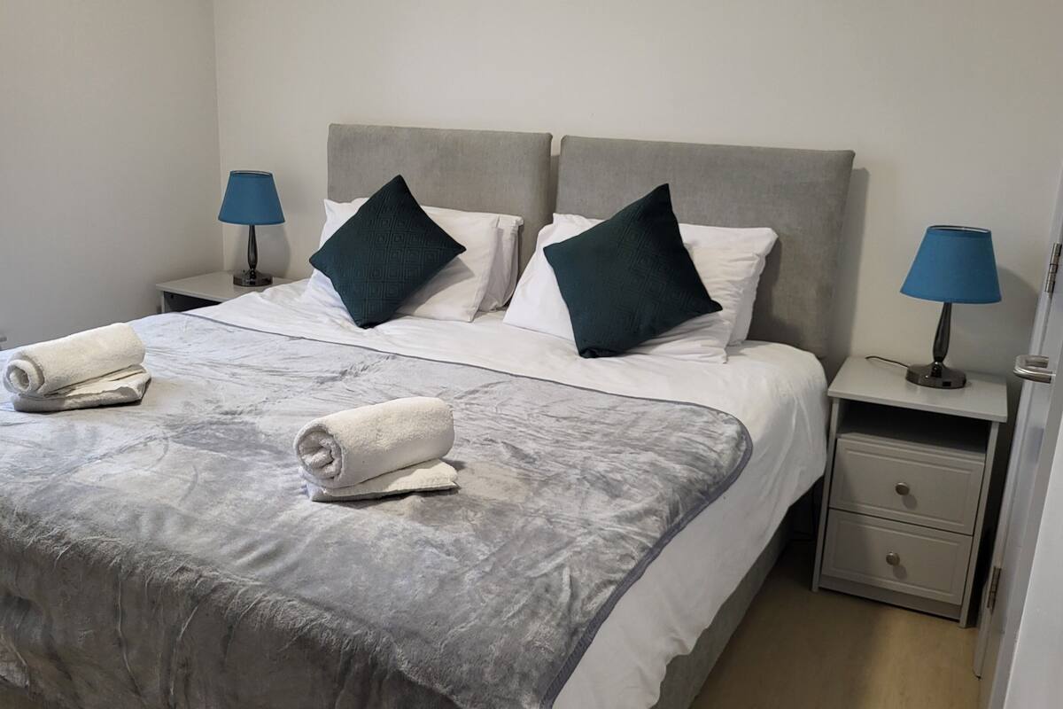 A neatly made bed with white and dark green pillows, two rolled towels, and a grey patterned bedspread. There are two bedside tables with lamps, a nightstand, and a drawer.