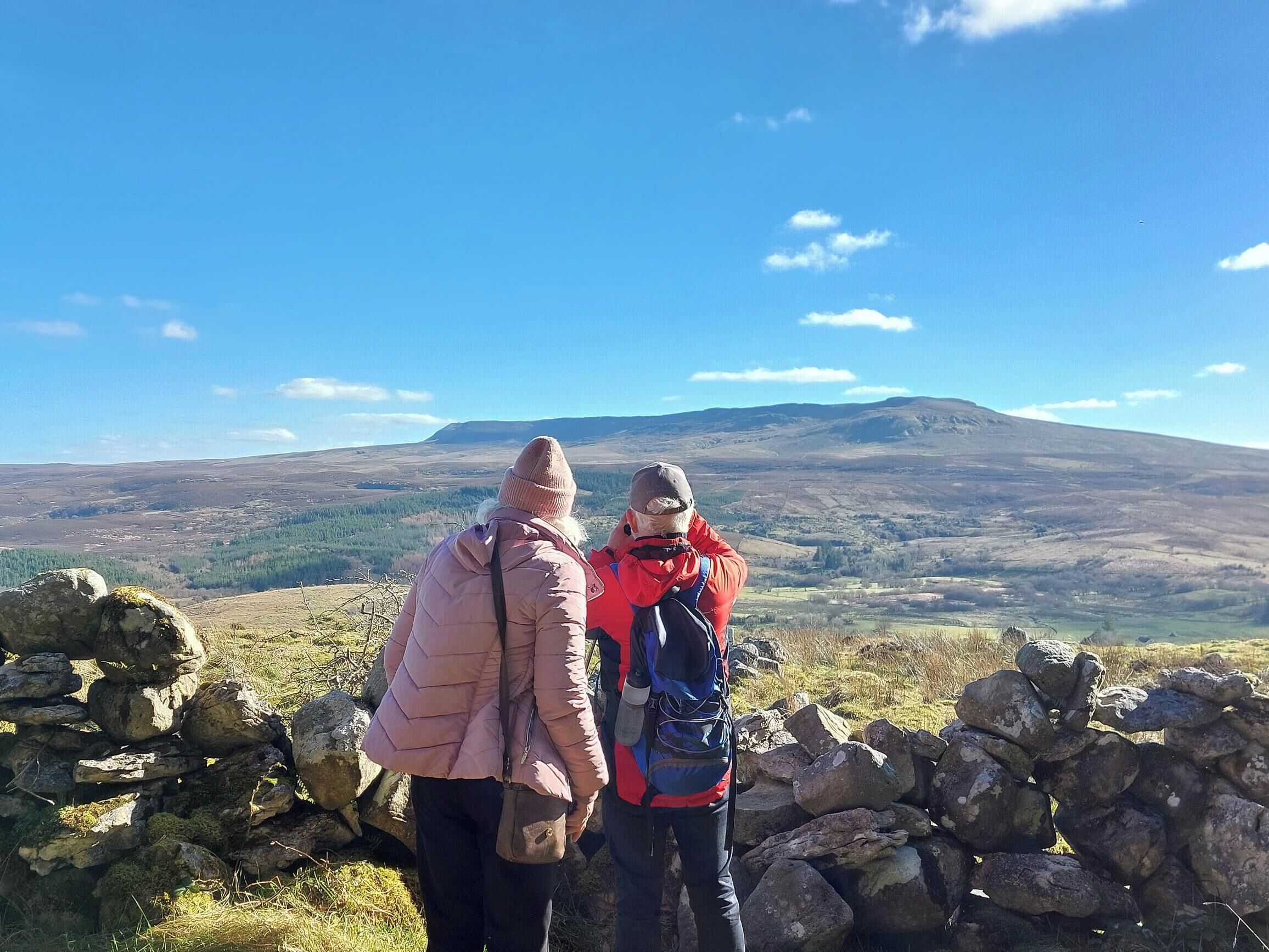 Two people standing by a stone wall overlooking a mountainous landscape.
