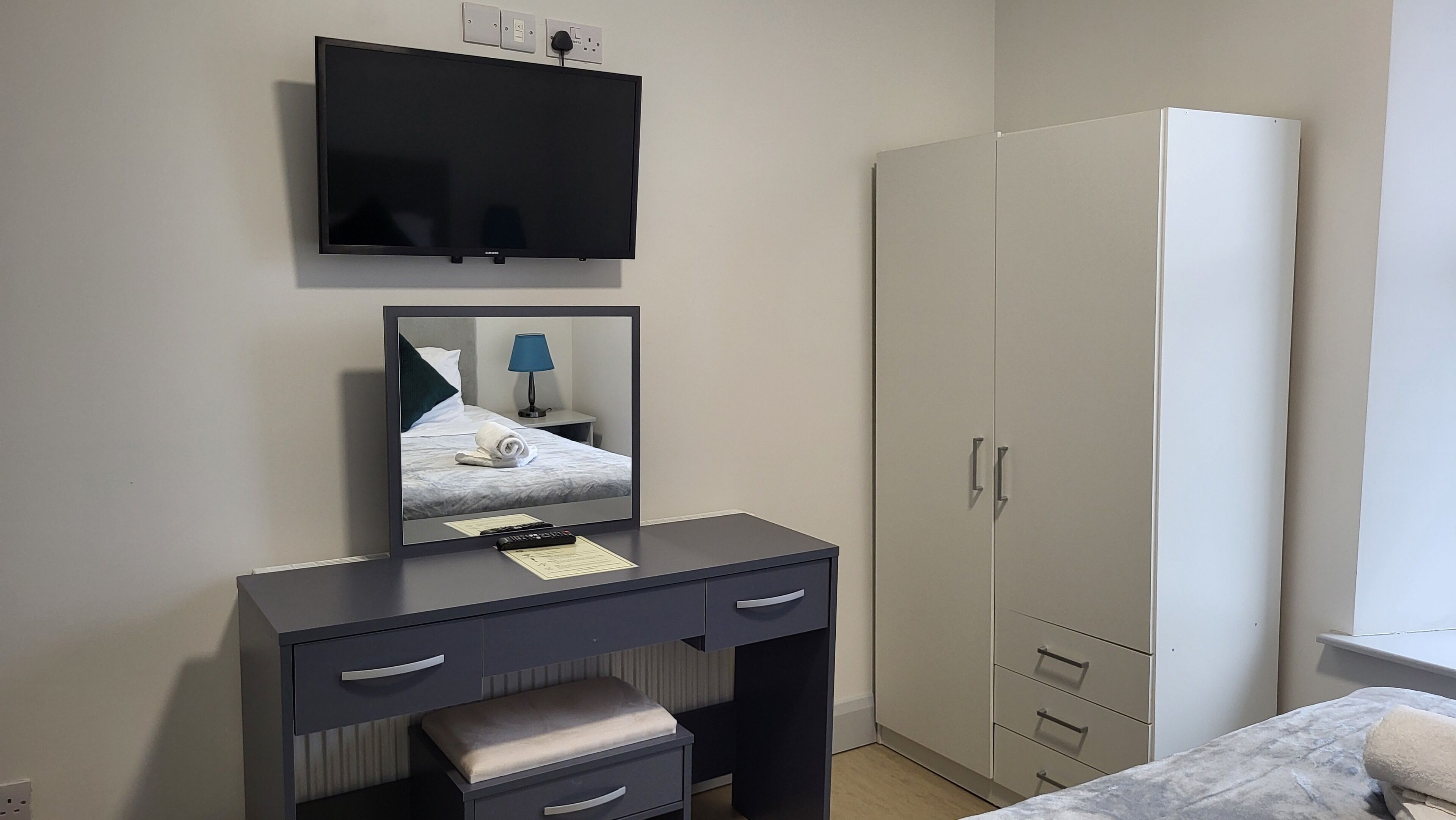 Een hotelkamer met een bed, een bureau met lades, een spiegel, een aan de muur bevestigde televisie en een kledingkast.