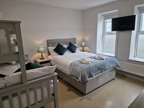 A bedroom with a bunk bed, a single bed, bedside lamps, a flat-screen TV, and a window. - Trivia House Accomodation (Furnaceland)