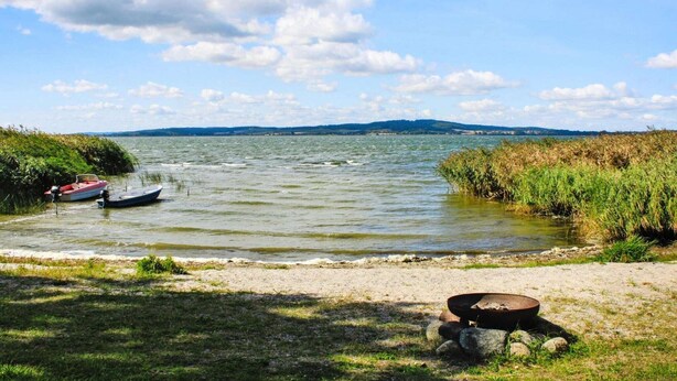 A lakeside scene with a boat docked near the shore, a fire pit, and a view of distant hills under a partly cloudy sky. - Ferienwohnung am Kummerower See, Sommersdorf (Sommersdorf)