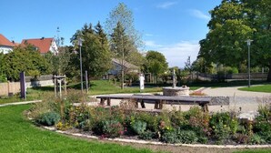 A park with a central stone bench, surrounded by flower beds and trees. - Ferienwohnung am Kummerower See, Sommersdorf (Sommersdorf)