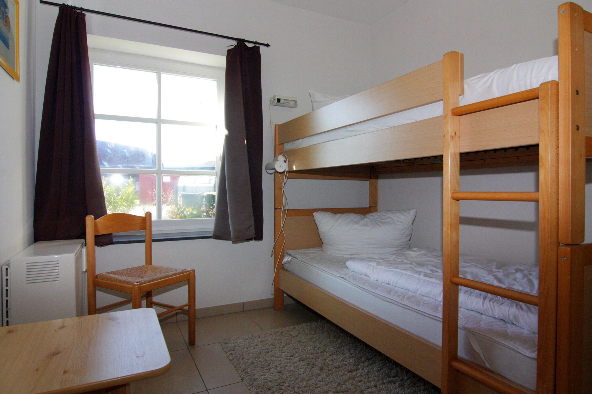 A bunk bed room with a wooden table and chair, a window with curtains, and a radiator.