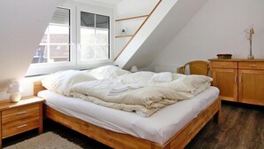 A bedroom with a sloped ceiling, a bed, a nightstand, a wooden dresser, and a window with blinds. - Ferienwohnung im Reihenhaus, Friedrichkoog-Spitze (Friedrichskoog-Spitze)