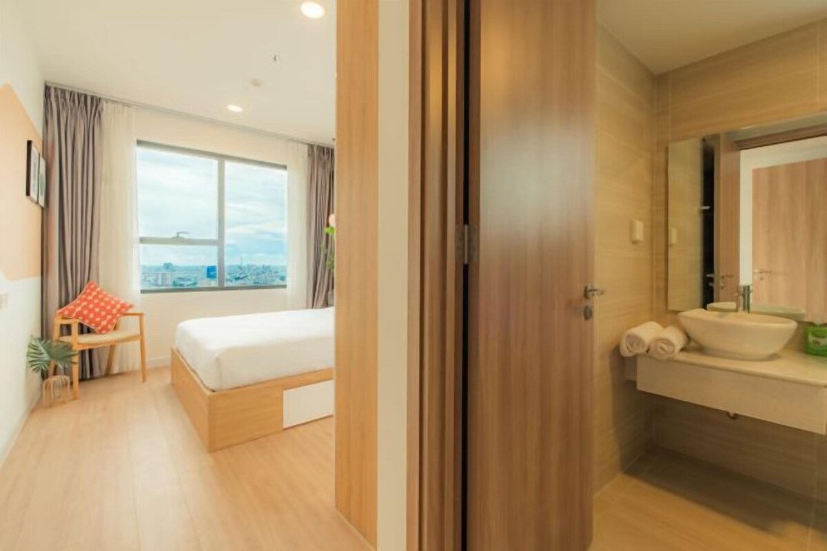 Une chambre d’hôtel avec un lit, une fenêtre donnant sur la ville, une salle de bain avec un lavabo et un miroir, et une porte.