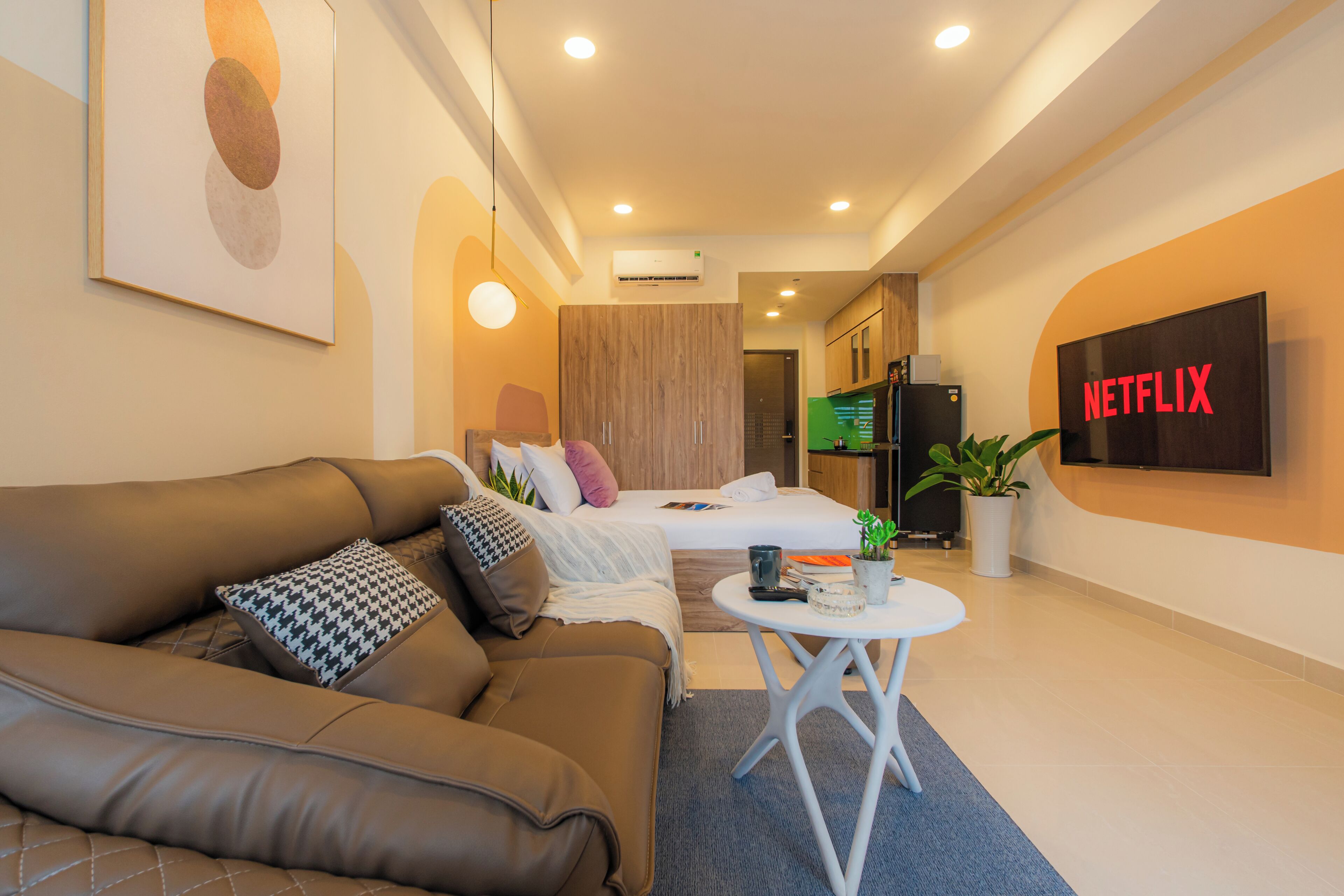 A modern hotel room with a brown leather sofa, a bed with a white and pink blanket, a small white table with a plant, and a wall-mounted TV displaying "NETFLIX".