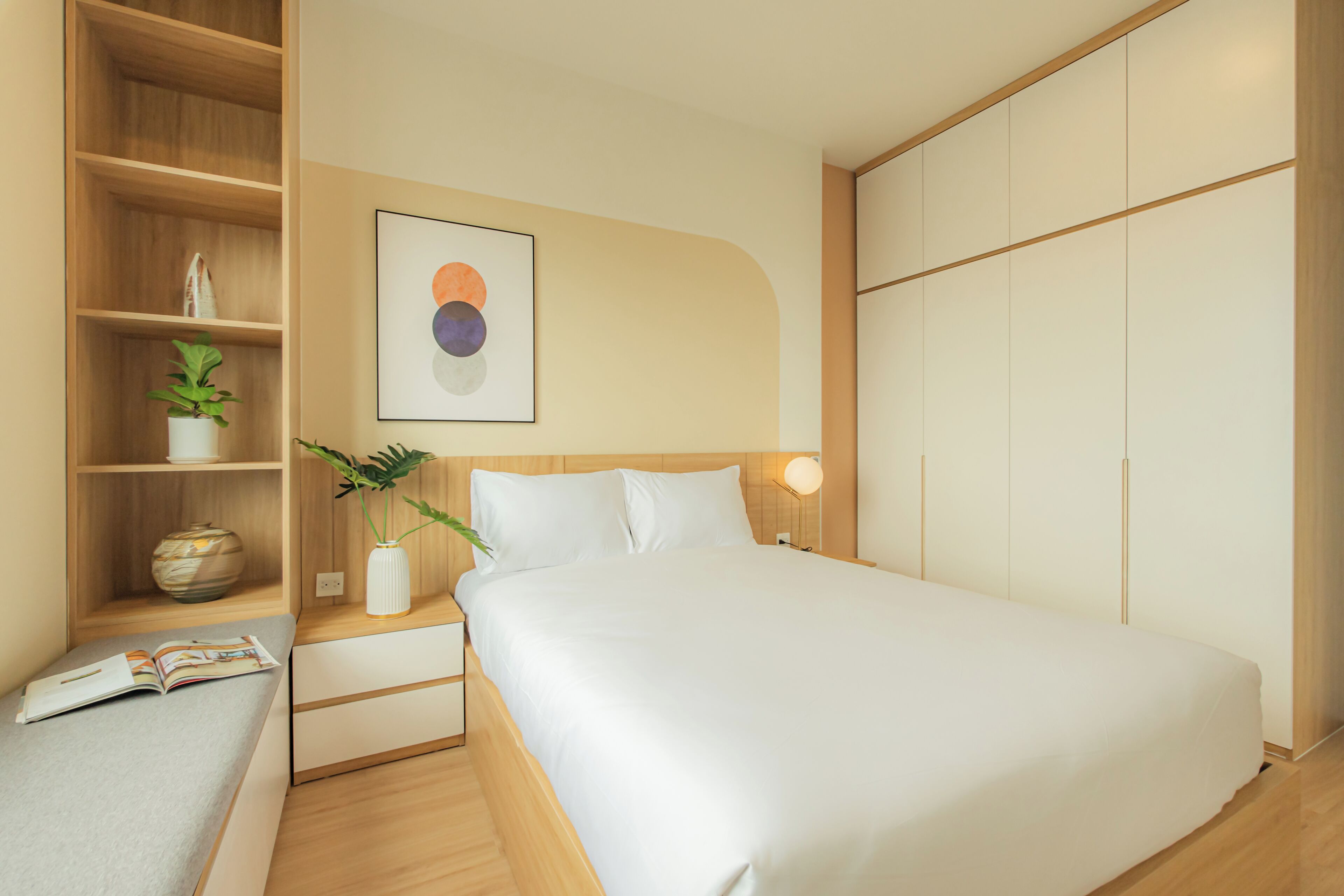 A modern bedroom with a large bed, a bedside table with a lamp, a shelf with decorative items, and a wall-mounted artwork.