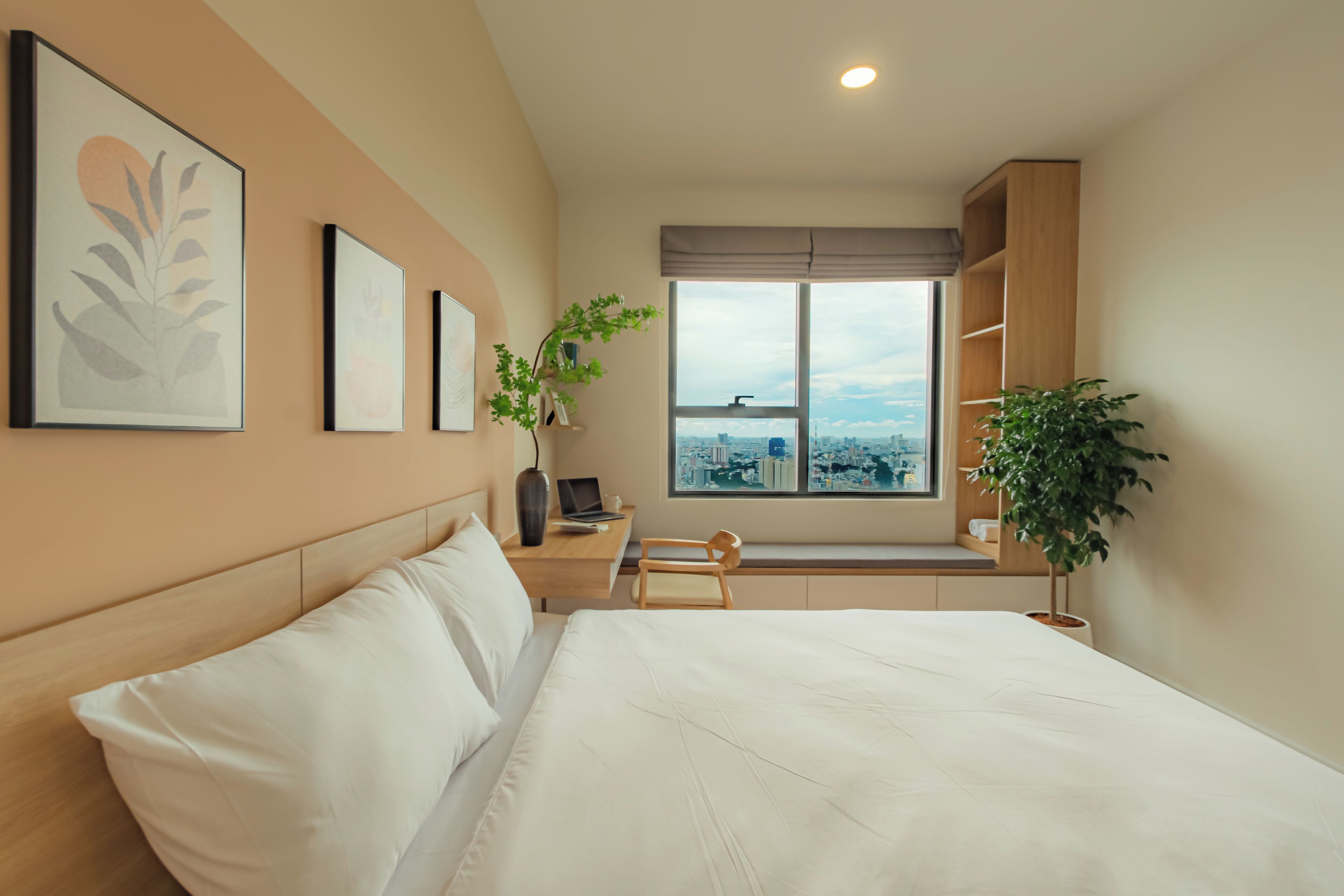 Une chambre d’hôtel moderne avec un grand lit, un bureau avec un ordinateur portable, une fenêtre donnant sur la ville et une plante en pot.