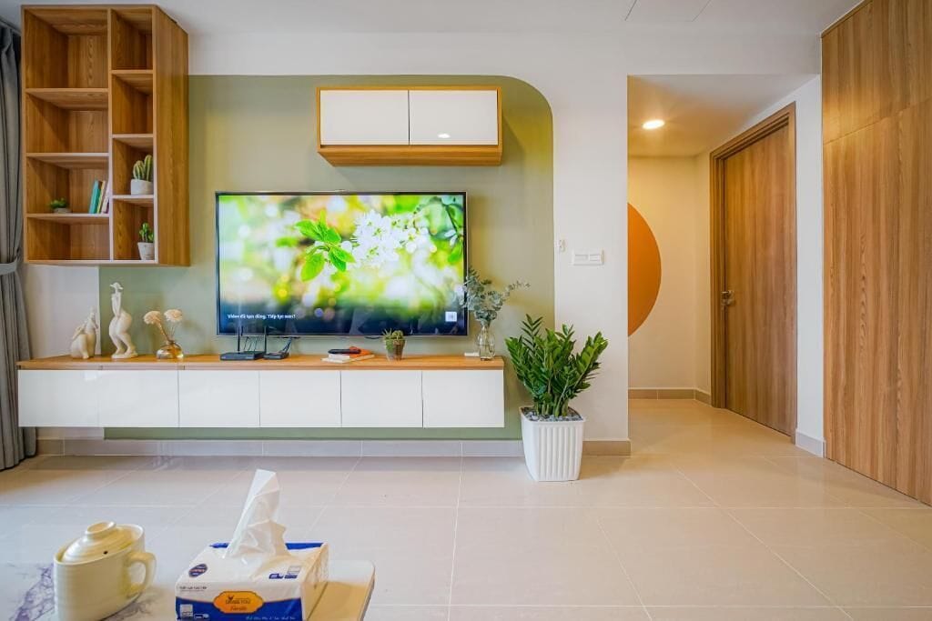A modern living room with a flat-screen TV mounted on a white cabinet, a wooden bookshelf, a potted plant, and a coffee table with a teapot and tissues.