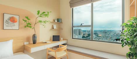 A modern bedroom with a bed, a desk with a laptop, a chair, a window with a city view, and a plant.
