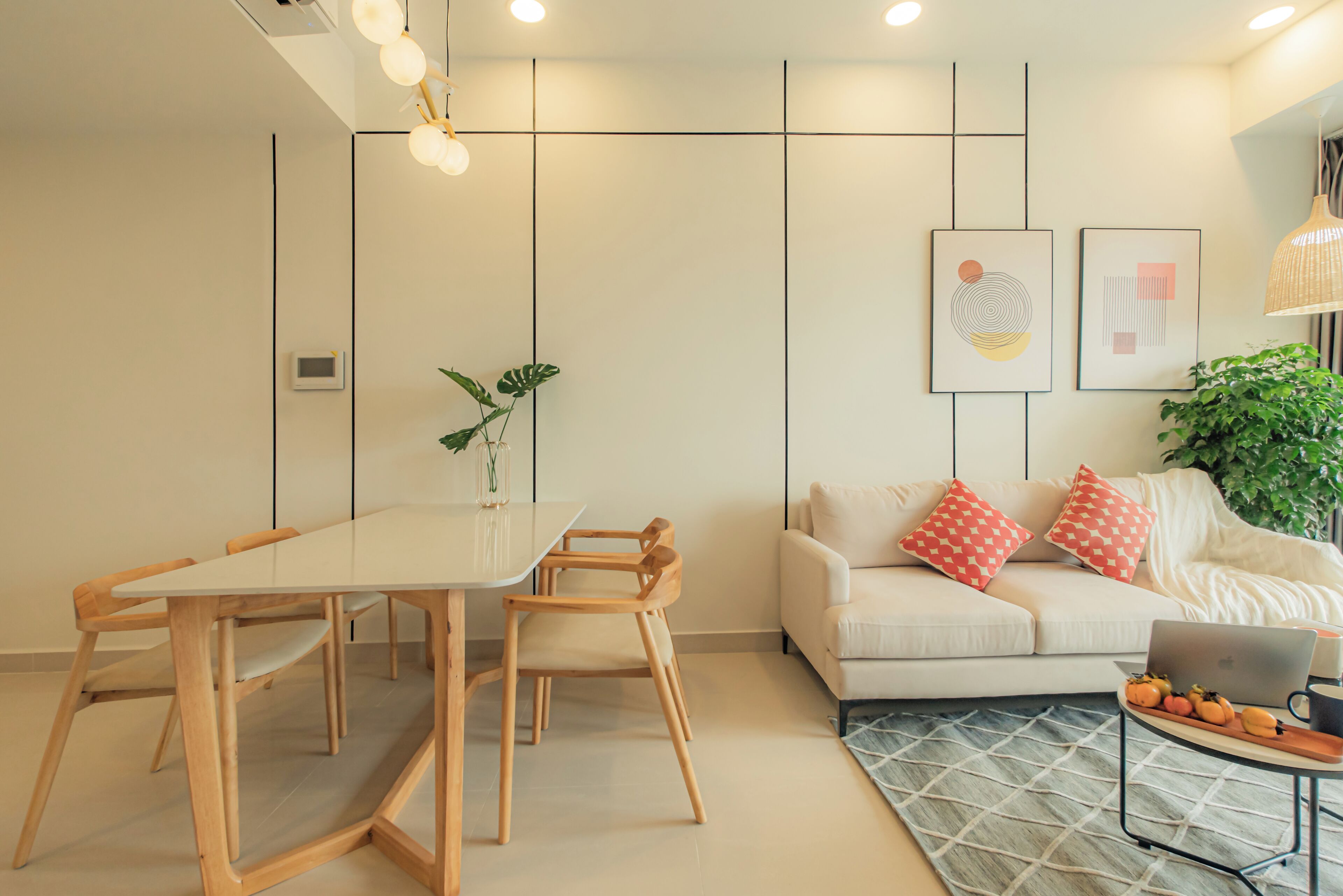 A modern living room with a dining area, a sofa with patterned cushions, a laptop on a side table, and a potted plant.