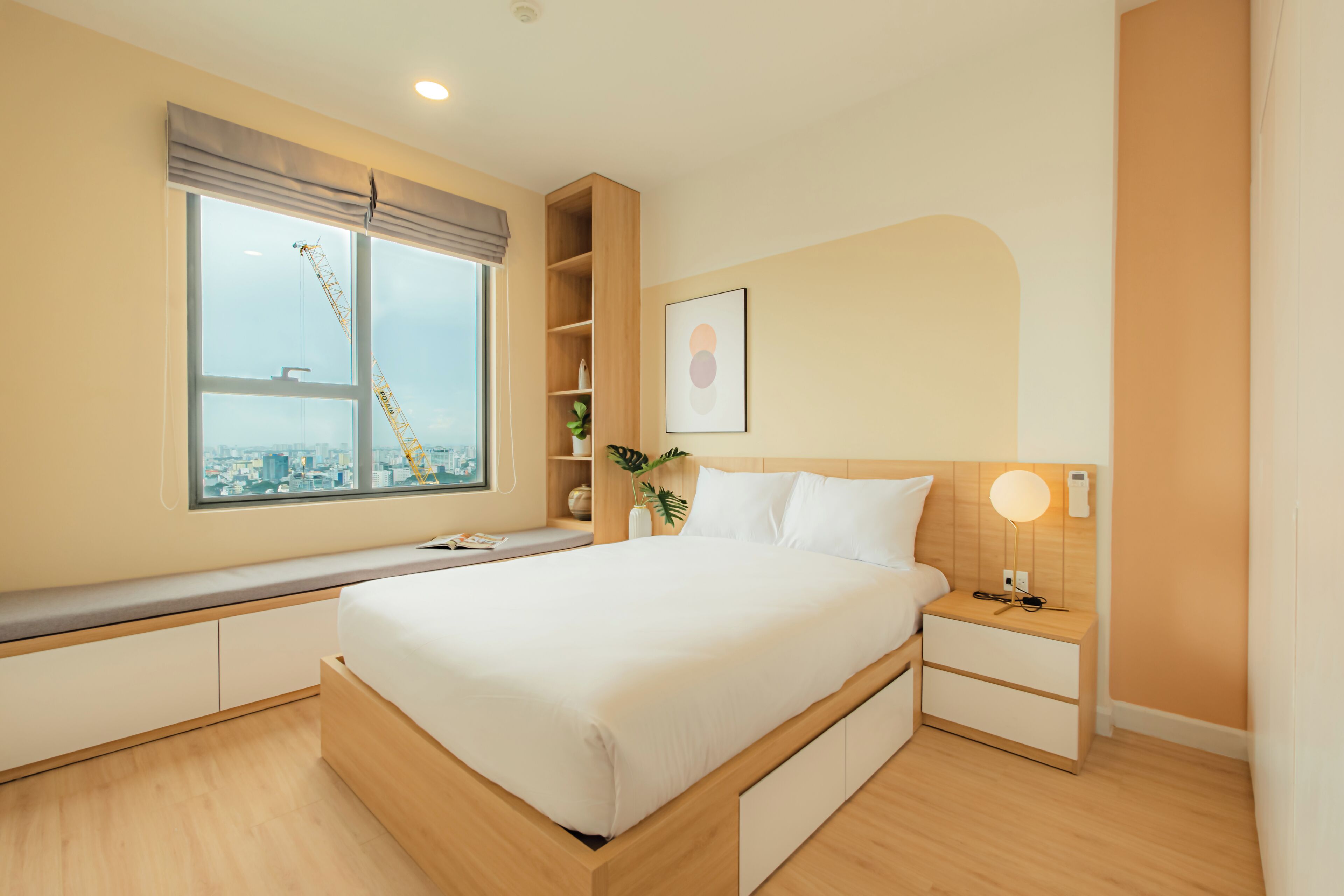 A modern bedroom with a large bed, built-in shelving, a bedside table with a lamp, and a window with a city view.