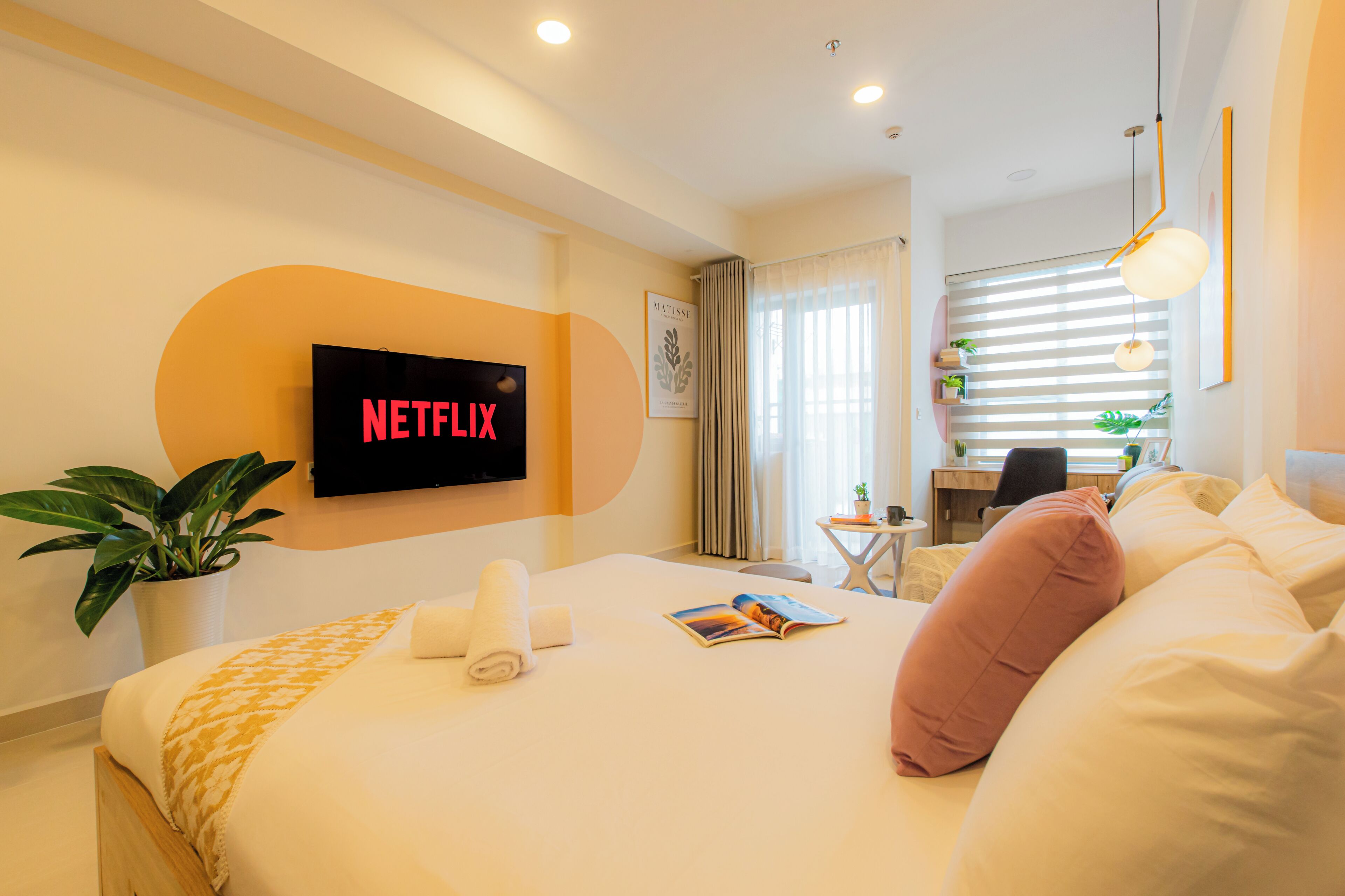 A hotel room with a bed, a desk, a chair, a potted plant, and a television displaying the Netflix logo.