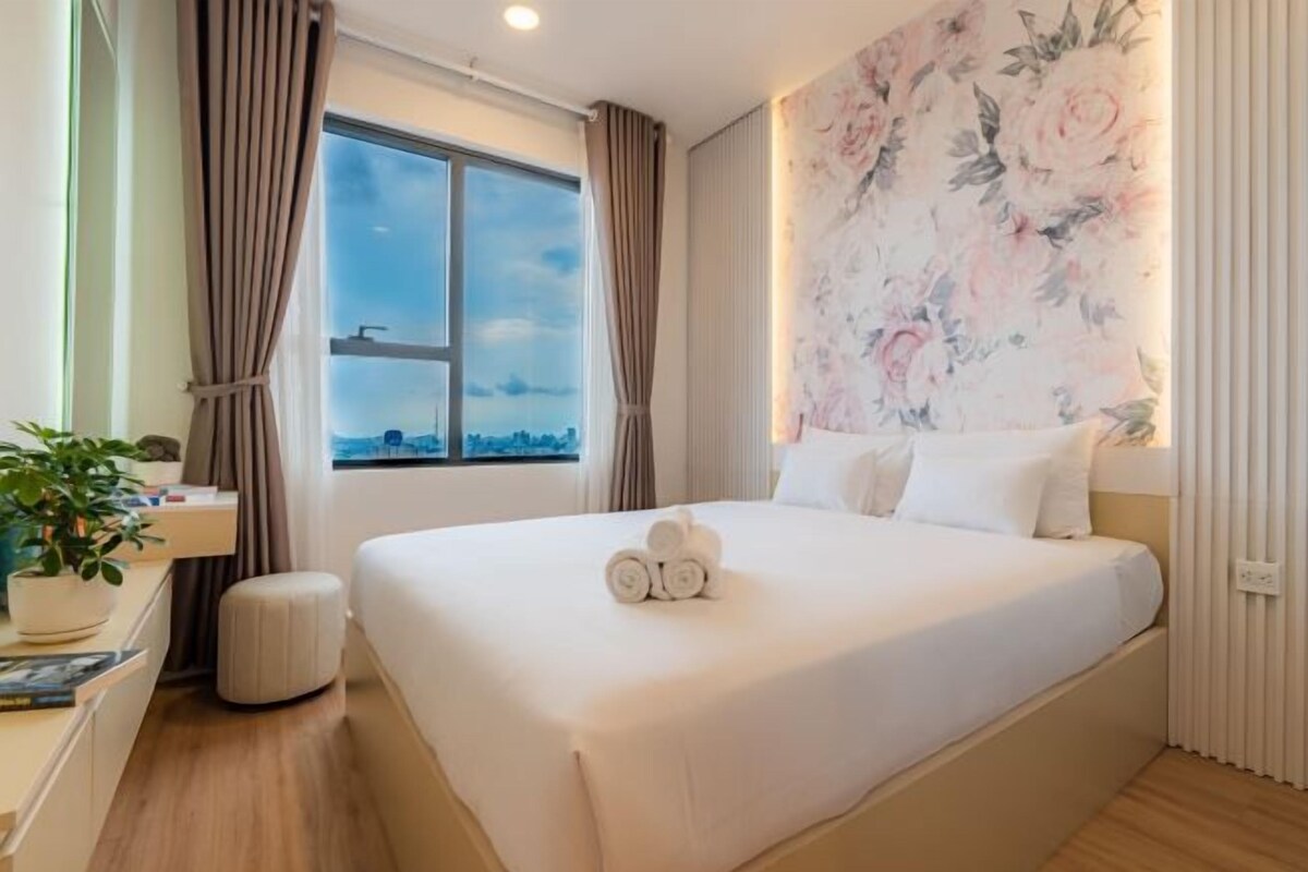 Une chambre d’hôtel avec un grand lit, une fenêtre avec des rideaux, une décoration murale florale et une petite table de chevet agrémentée d’une plante.