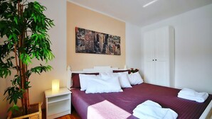 A neatly made bed with white linens, a cityscape painting above it, a bamboo plant, and a white nightstand with a lamp.