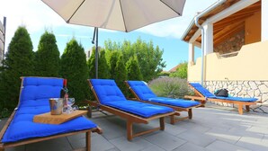 A patio with three sun loungers, each with a blue cushion and a wooden table in front.