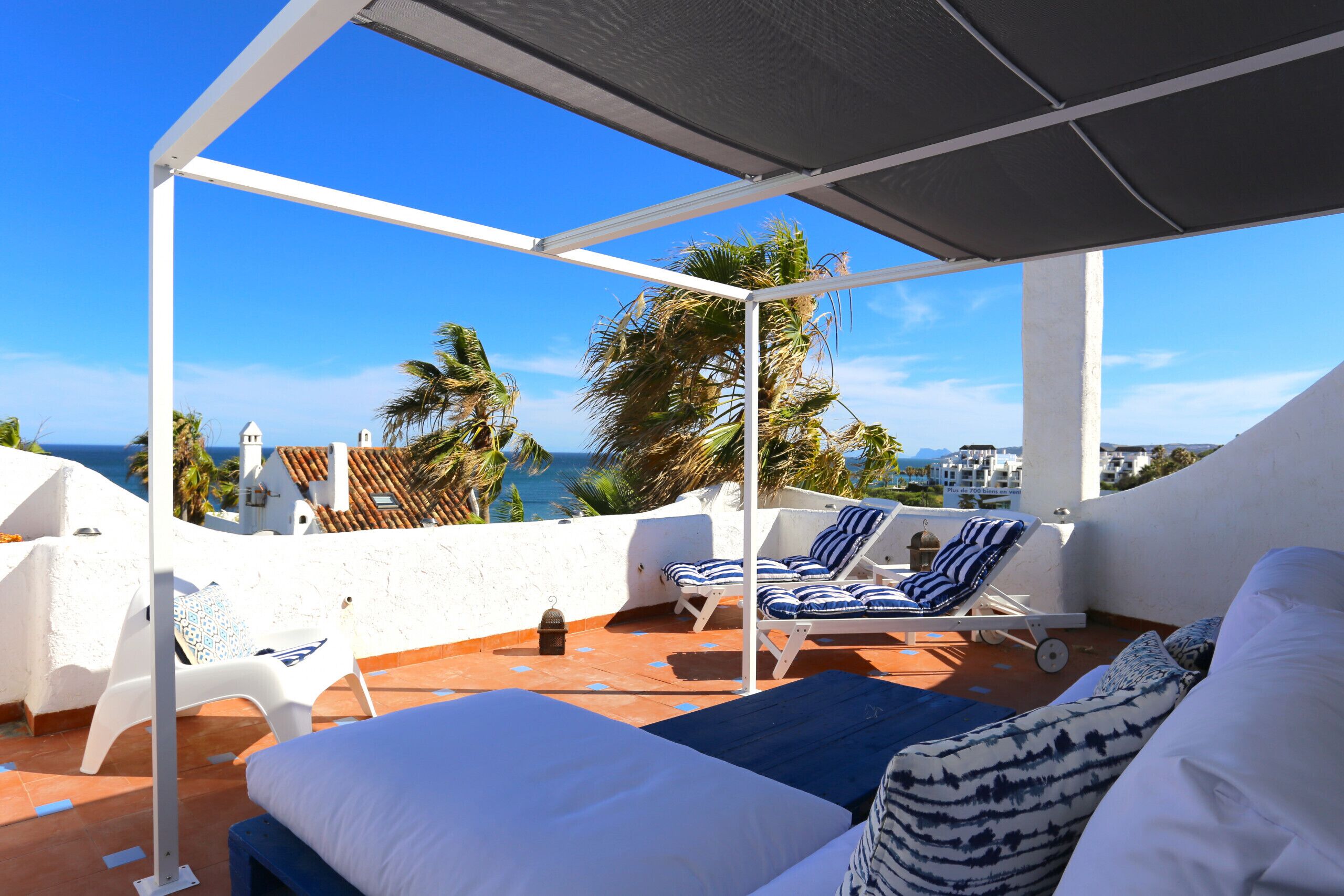 A rooftop terrace with white walls, blue-striped lounge chairs, and a bed with patterned pillows.