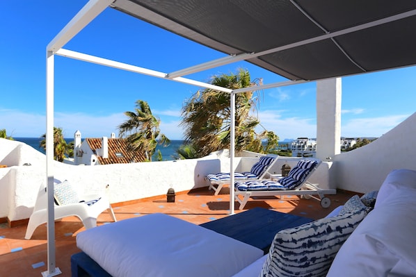 Terraza en la azotea con paredes blancas, sillones de jardín azul y blanco a rayas, y una cama con almohadones estampados.