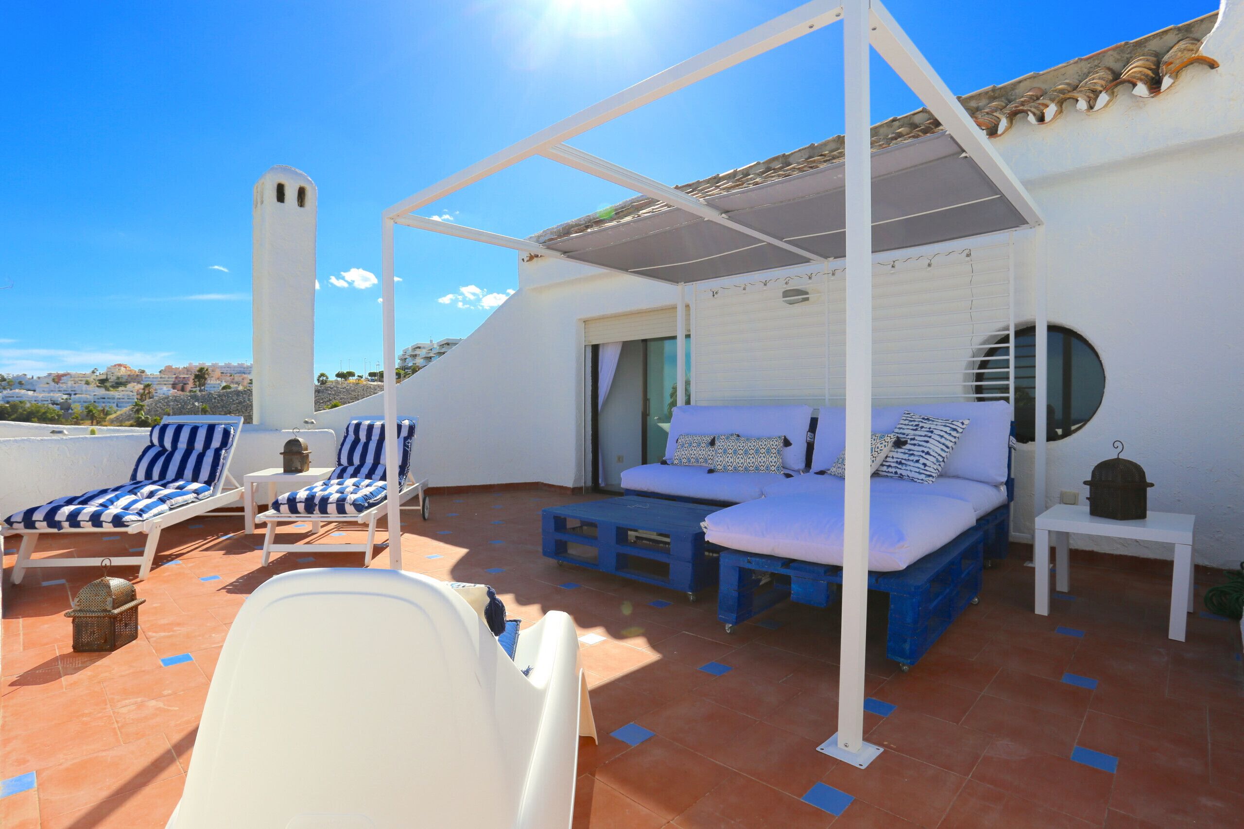 A rooftop terrace with a white pergola, blue and white striped lounge chairs, a blue and white sofa, and a small table with a pot.