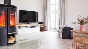 Ein modernes Wohnzimmer mit einem schwarzen Kaminofen, einem Flachbildfernseher, einem Glastisch, einem runden Holztisch mit einem Stuhl, einem dunklen Sofa und einem Fenster mit Vorhängen.