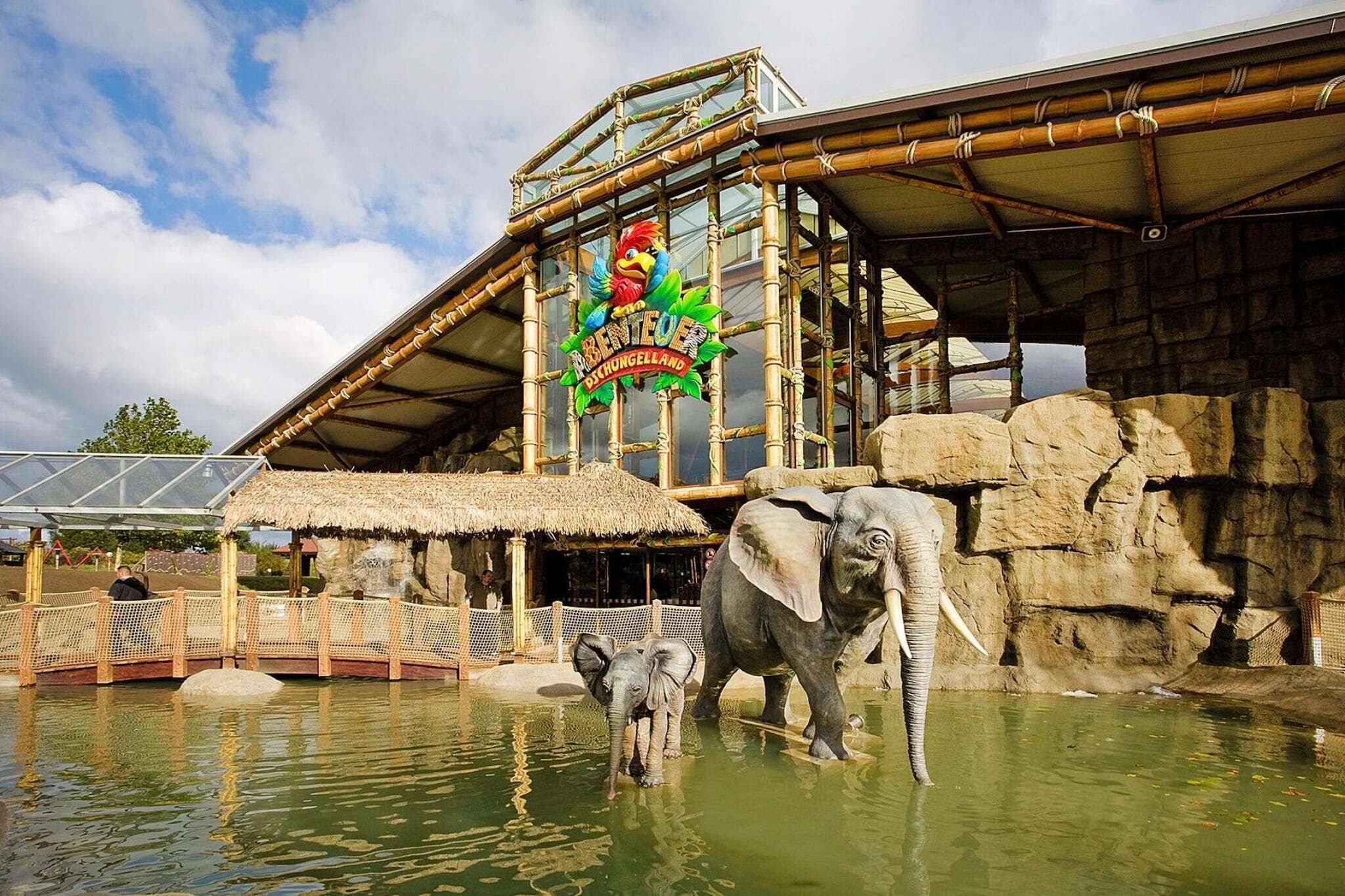 A zoo enclosure with an elephant and a baby elephant, a thatched-roof structure, and a large sign with a bird and text.