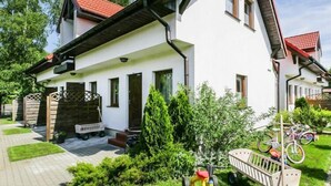 Ein zweistöckiges Haus mit rotem Ziegeldach, einer Veranda und einem kleinen Garten mit Rasen und Blumentöpfen.