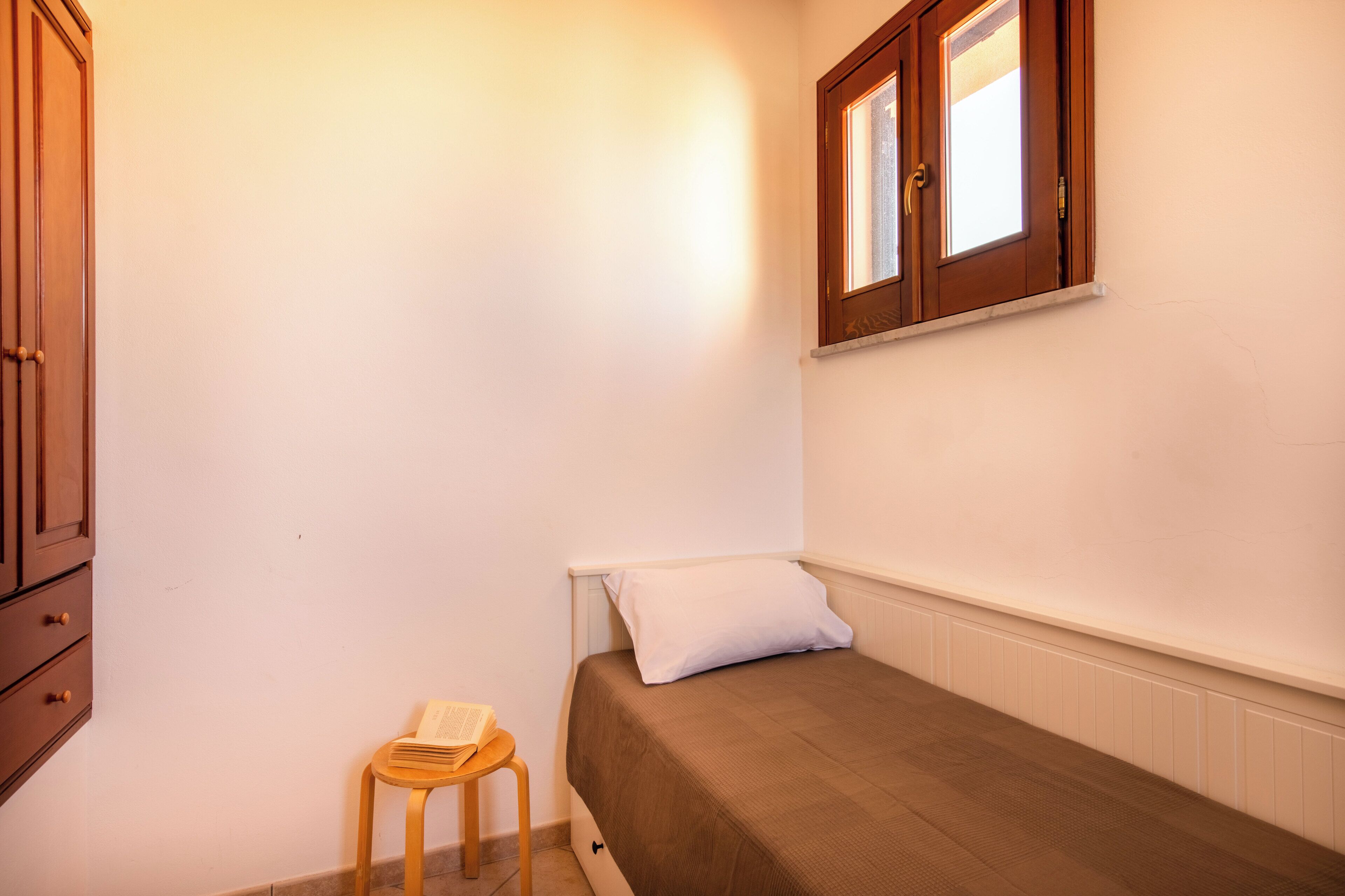 Habitación pequeña con cama individual, un armario de madera, un taburete con un libro y una ventana con marcos de madera.