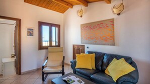 A living room with a leather sofa, a chair, a coffee table with a magazine, a window with a view, and a wall with a textured artwork. - Terrazza Mandralisca by Wonderful Italy (Cefalù)