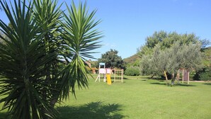 A grassy outdoor area with a palm tree, a playground, and a building with a sign. - Apartment in Elba near Pebble Beach (Rio nell'Elba)
