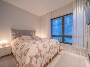 A bedroom with a bed, nightstands, a window with blinds, and a view of trees.