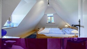 Um quarto com teto inclinado, uma cama com colchão branco e roupa de cama azul, uma janela pequena com vista e uma mesa de cabeceira com abajur.